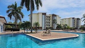Ein Swimmingpool mit Liegestühlen, Palmen und modernen Gebäuden im Hintergrund.