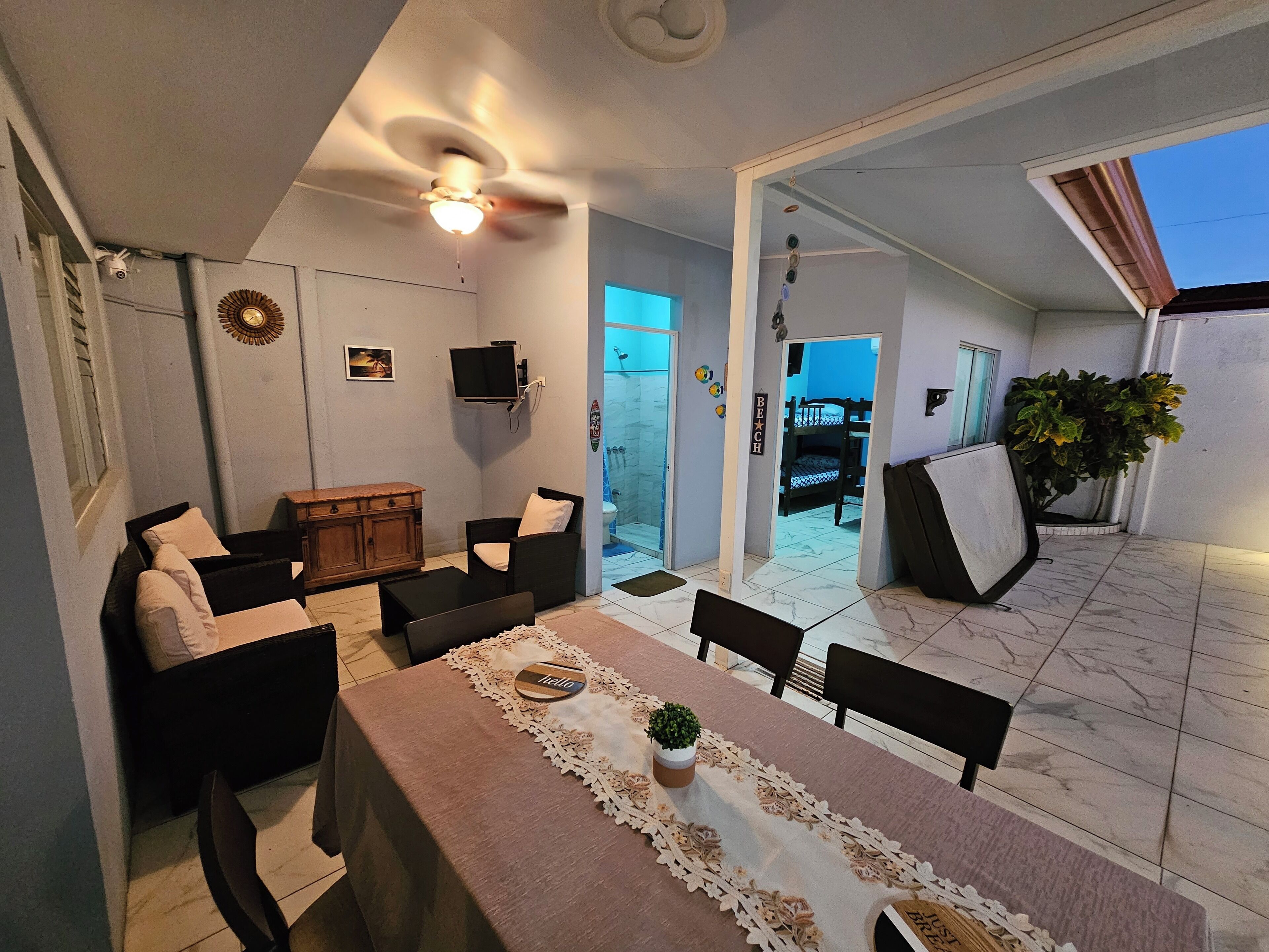 Un espace intérieur accueillant avec une table à manger, des chaises, un canapé et un ventilateur de plafond. On trouve une petite plante sur la table et un téléviseur fixé au mur.