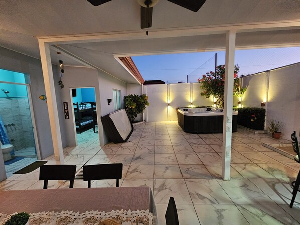 Un patio exterior techado con jacuzzi, zona de estar y un baño con paredes de vidrio.