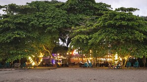 Eine Strandlocation mit Außenbestuhlung unter Bäumen, Lichterketten und einer Bühne.