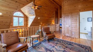 A spacious wooden cabin interior with a high ceiling, a ceiling fan, a large window, a fireplace, a wooden door, and a bathroom with a toilet and sink. - Morris Cabin- Cedar log cabin with hot tub, dog friendly, mountain views (Boone)