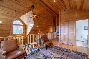 A spacious wooden cabin interior with a high ceiling, a ceiling fan, a large window, a fireplace, a wooden door, and a bathroom with a toilet and sink.