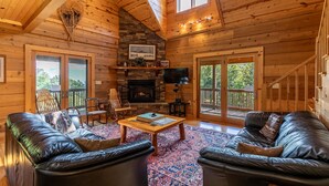 A cozy cabin interior with a stone fireplace, leather sofas, a wooden coffee table, and a TV mounted on the wall. - Morris Cabin- Cedar log cabin with hot tub, dog friendly, mountain views (Boone)