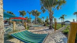 Un hamac installé sur une plage de sable, avec des palmiers et un ciel bleu clair en arrière-plan.