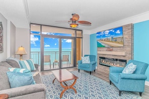 A living room with a sofa, armchairs, a coffee table, a fireplace, and a balcony with a view of the ocean.