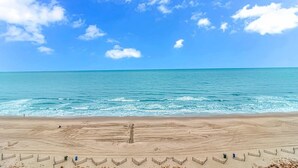 Een strand met een heldere lucht, een zandgebied met hekken, en de oceaan op de achtergrond.