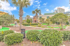 A golf course with manicured hedges, palm trees, and a building with a green roof in the background.