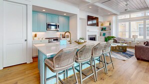 Une cuisine moderne avec des meubles de couleur turquoise, un îlot central blanc doté de tabourets de bar et un coin repas comprenant un canapé et une table basse.