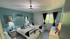 Une chambre avec deux lits, un ventilateur de plafond, une fenêtre avec des rideaux, une table de chevet avec une lampe et une commode.