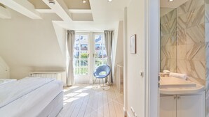 Et moderne soveværelse med en stor seng, en stol, et badeværelse med badekar og udsigt til bygninger gennem et vindue.