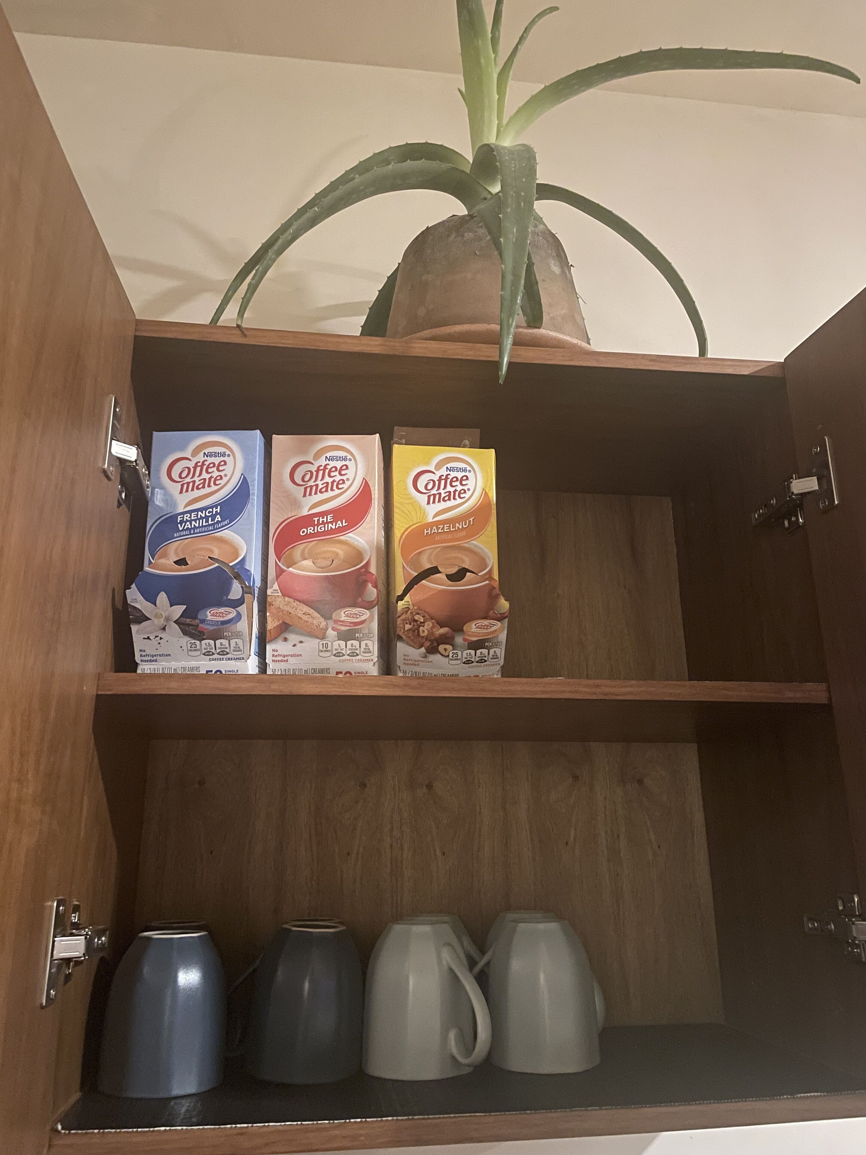 Un meuble en bois avec trois boîtes de Nestlé Coffee Mate sur l’étagère supérieure et trois tasses à café sur l’étagère inférieure.