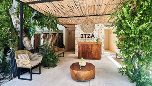 Un espace extérieur abrité, aménagé avec un banc en osier, une table ronde en bois et un mur de pierre sur lequel est inscrit le mot « ITZÁ ».