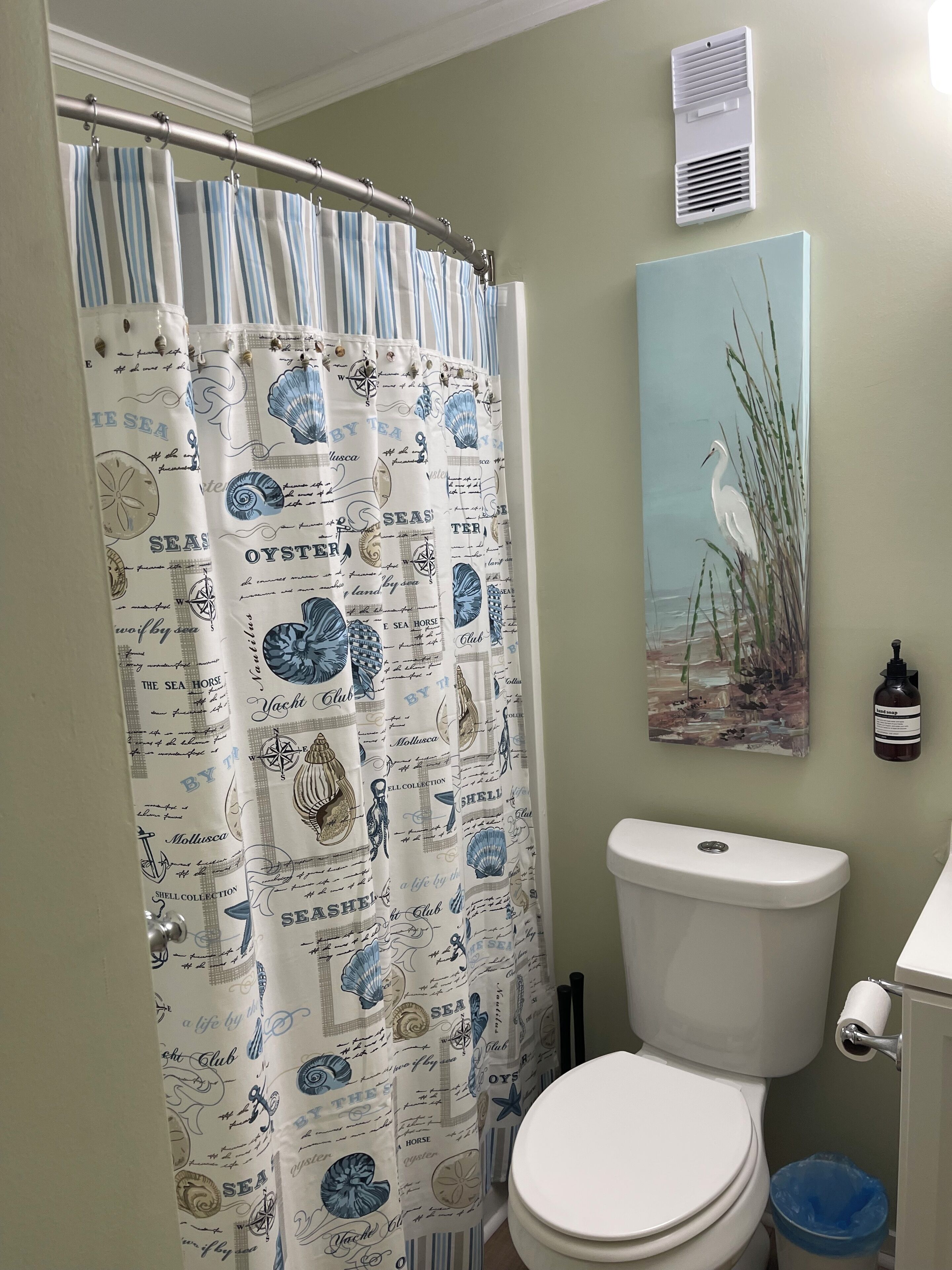 Un baño con cortina de ducha con motivos náuticos, un inodoro, una rejilla de ventilación en la pared y un cuadro de un pájaro.