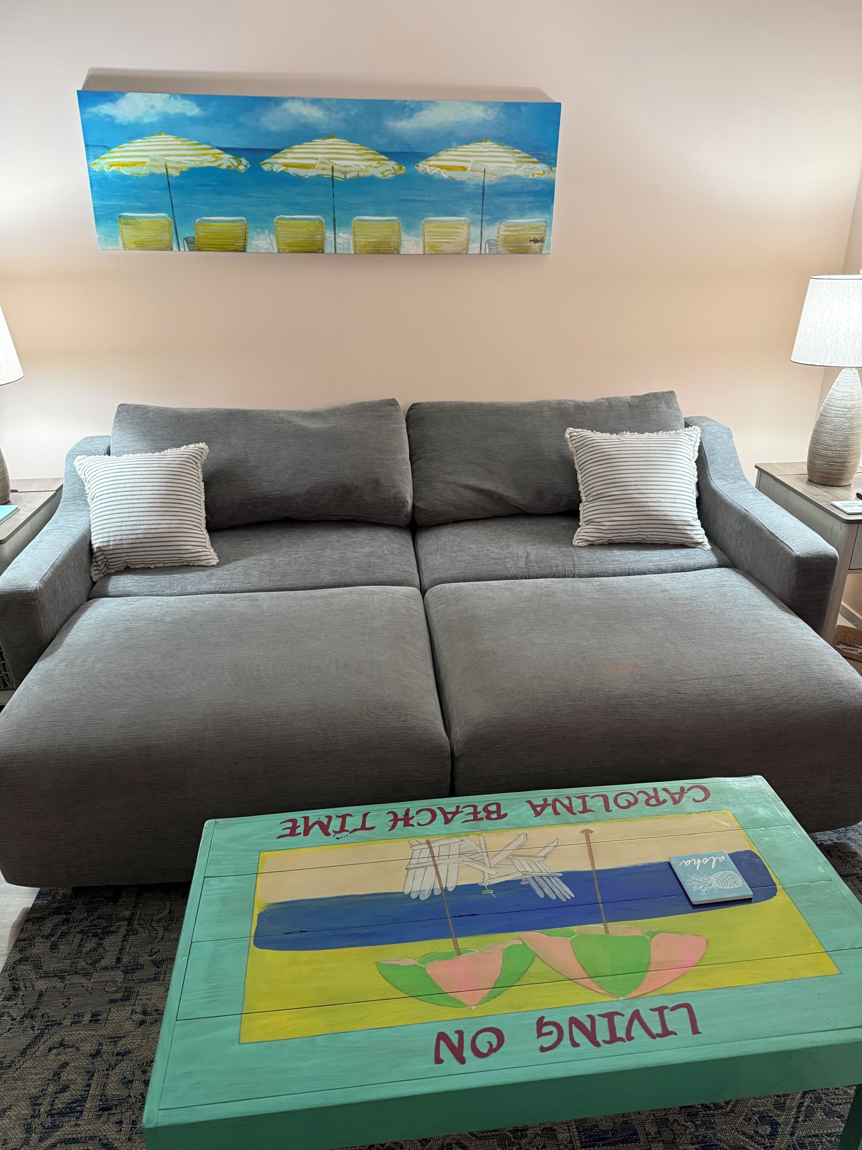 Un sofá gris con almohadones a rayas, una mesa de centro colorida con diseño de pájaro y frutas, y un cuadro de sombrillas de playa arriba.
