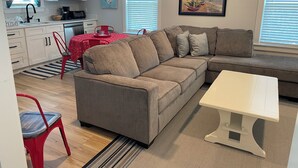 Una sala moderna con un sofá modular, una mesa de comedor con mantel rojo, una zona de cocina con armarios blancos y un cuadro en la pared.