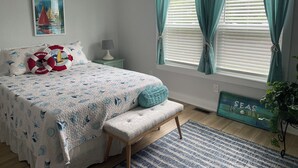 Un dormitorio con cama, un banco, una mesita de noche, una lámpara, una ventana con cortinas, un cuadro en la pared y una planta.