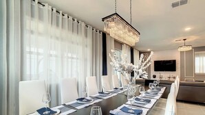 Een moderne eetruimte met een lange tafel voor zes personen, witte stoelen en een kroonluchter.