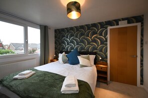 A hotel room with a large bed, a window with a view, a wooden door, and a patterned wallpaper.