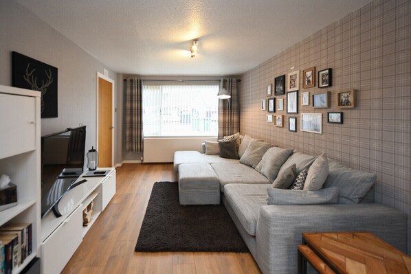 A modern living room with a grey sofa, a black rug, and a wooden bench. There are framed pictures on the wall and a window with curtains.