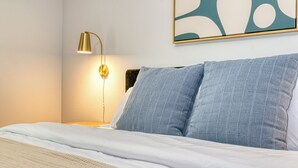 Una cama bien hecha con sábanas blancas, un cojín azul con textura y una lámpara dorada sobre una mesita de noche.