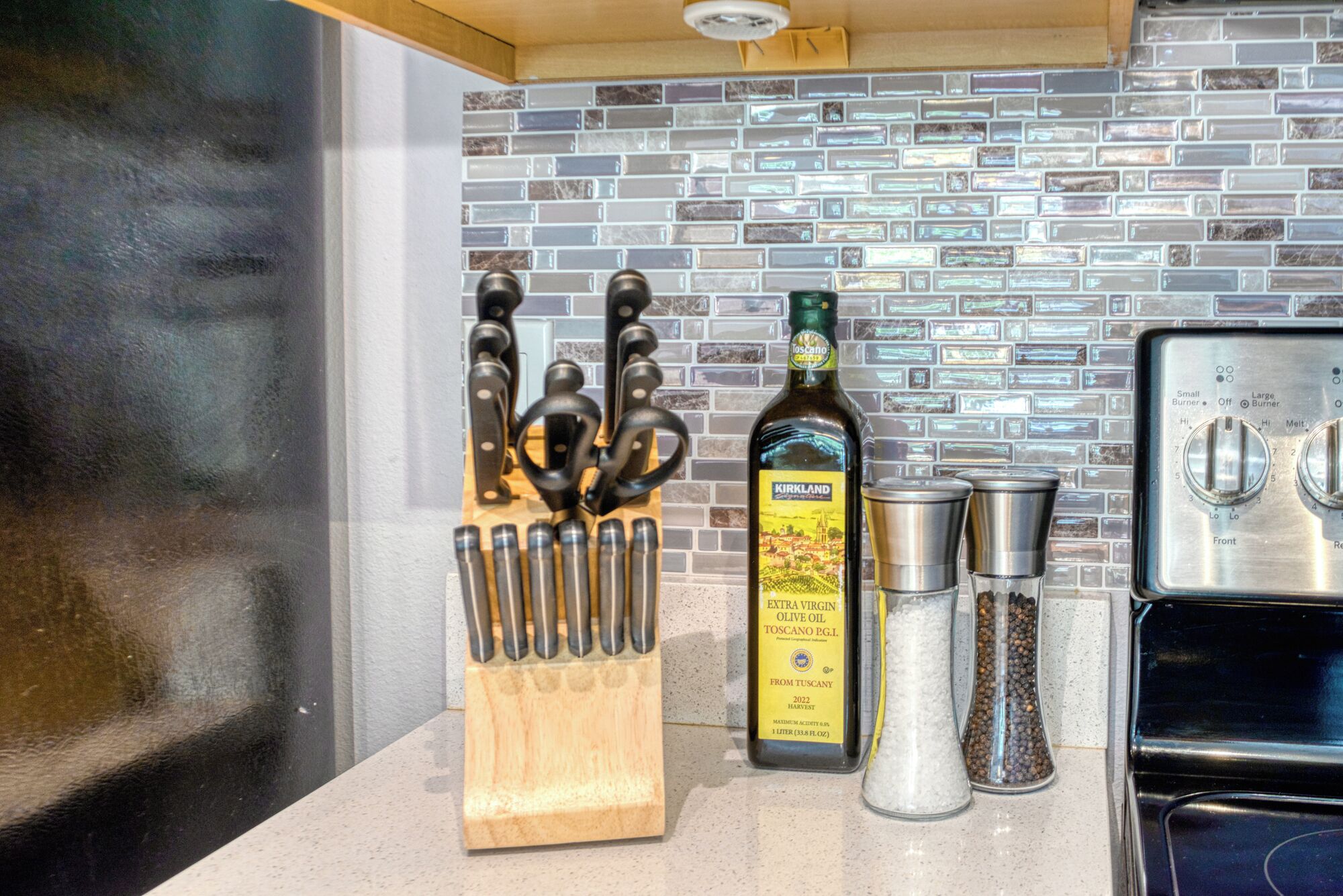 A kitchen counter with a knife block, a bottle of Kirkland Extra Virgin Olive Oil, salt and pepper shakers, and a stove.