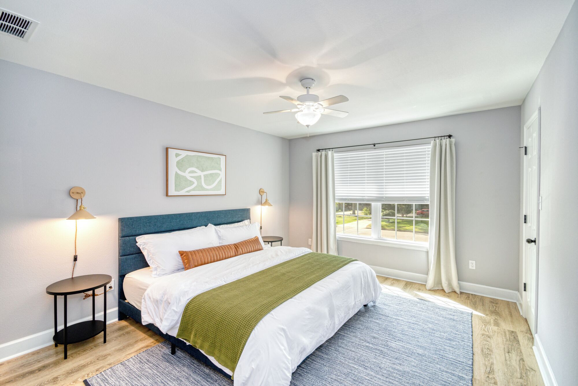 A bedroom with a bed, two bedside tables, a ceiling fan, a window with curtains, and a wall-mounted artwork.