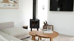 Un salon comprenant un canapé, une table basse ronde en bois, un poêle à bois noir et un téléviseur à écran plat fixé au mur.