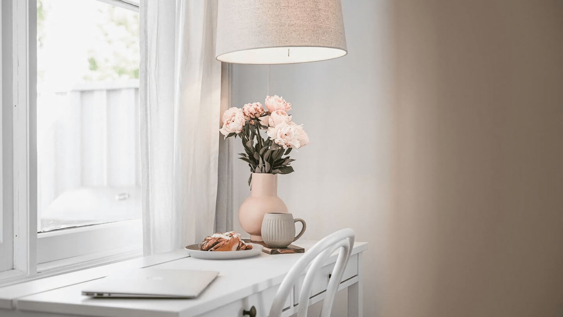 Un bureau blanc avec un ordinateur portable, une assiette de biscuits, une tasse et un vase rempli de fleurs.