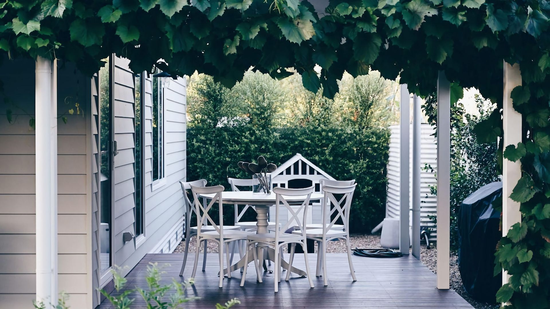 A covered patio with a table and chairs, surrounded by a white fence and greenery.