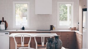 Een keuken met witte kastjes, een wit aanrecht en twee ramen. Er staan twee witte stoelen bij een tafel met twee metalen kannen.