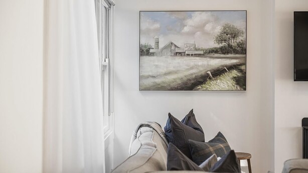 Een kamer met een schilderij van een landelijke omgeving, een aan de muur bevestigde televisie en een raam met gordijnen.