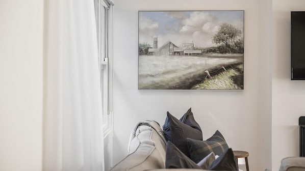 A room with a painting of a rural scene, a television mounted on the wall, and a window with curtains.