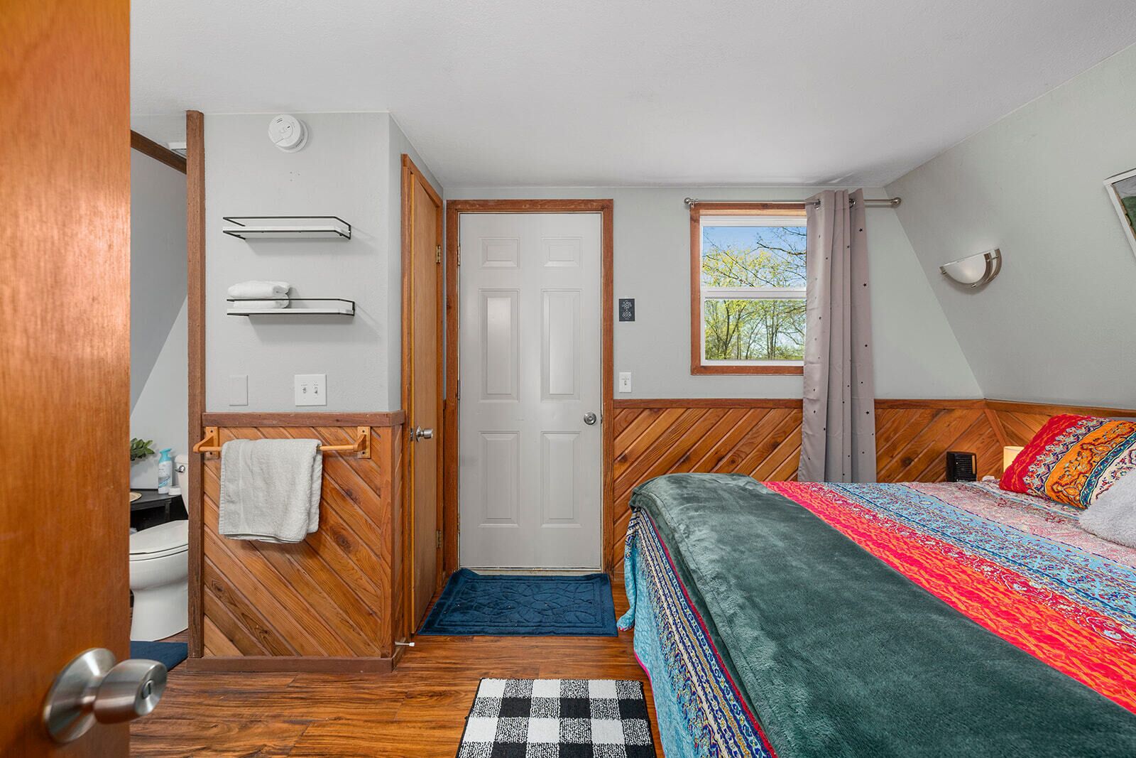 A compact bedroom with a bed, a window with a view, a door, and a small bathroom area.