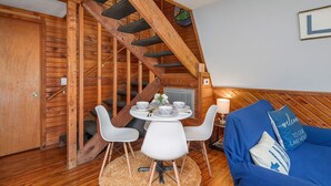 Une pièce chaleureuse avec un escalier en bois, un coin repas avec des chaises blanches et une table dressée, ainsi qu’un canapé bleu agrémenté d’un coussin sur lequel est inscrit : « Bienvenue dans notre maison au bord du lac ».