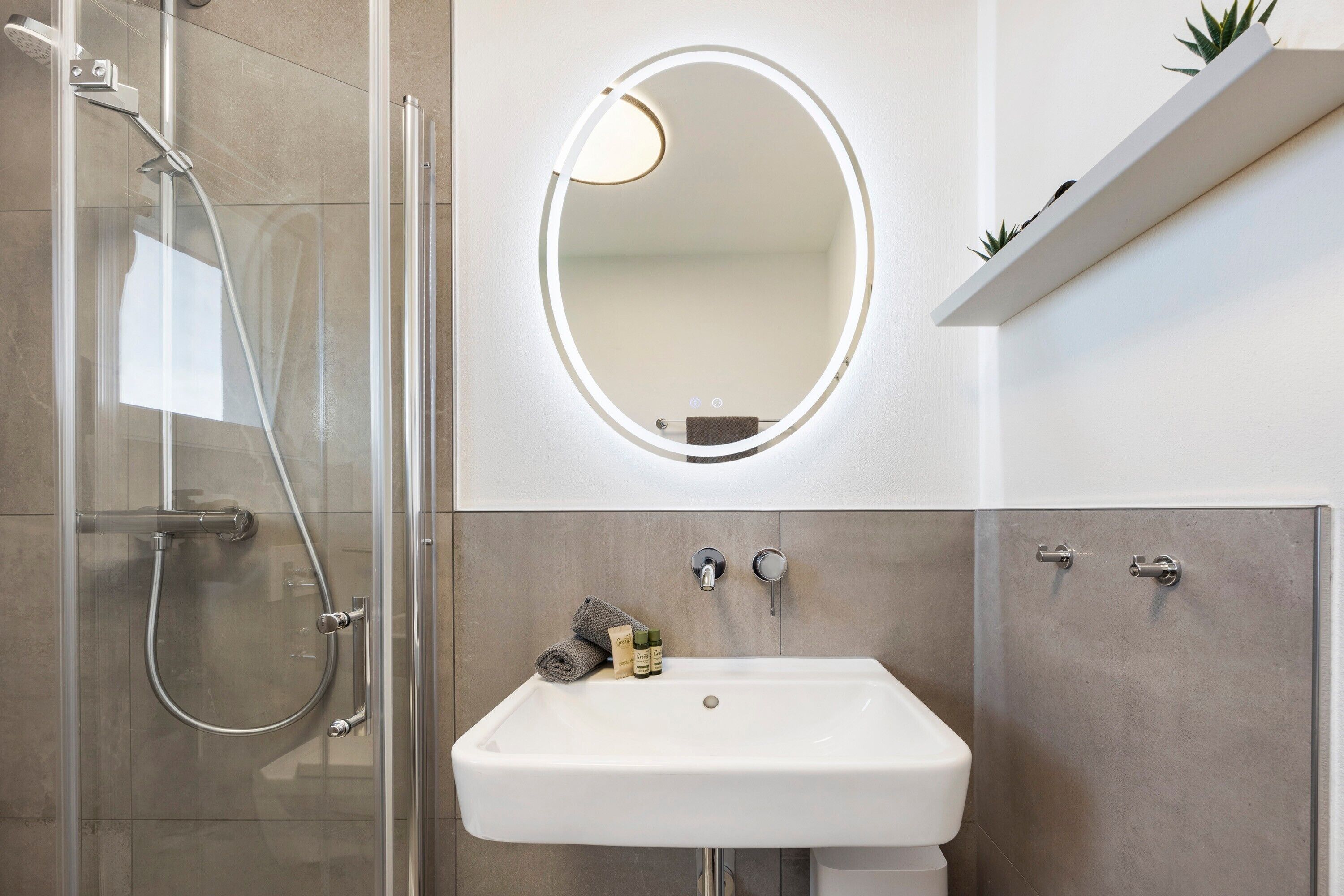 A bathroom with a glass-enclosed shower, a white sink, a round mirror with a light, and a shelf with plants.