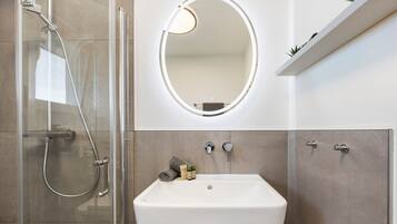 A bathroom with a glass-enclosed shower, a white sink, a round mirror with a light, and a shelf with plants.