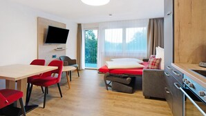 A modern hotel room with a wooden floor, a flat-screen TV mounted on the wall, a dining table with red chairs, a sofa bed with a red cushion, and a kitchenette with a stove. - Ferienwohnung Rothirsch, 45 qm, 1 Schlafzimmer, 1 Schlafsofa, max. 4 Personen (Höchenschwand)
