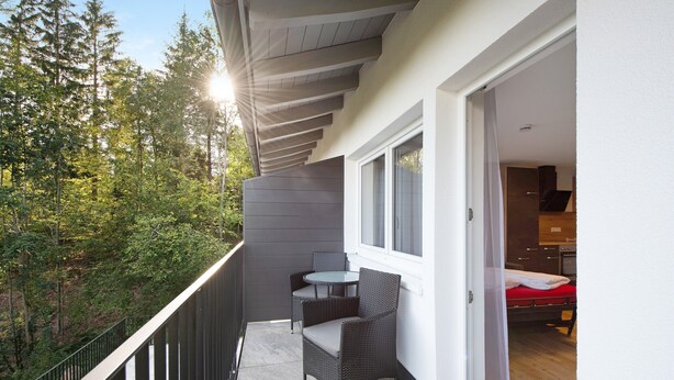A balcony with a table and chairs, a view of trees, and a glimpse of a room with a bed and a kitchen area. - Ferienwohnung Rothirsch, 45 qm, 1 Schlafzimmer, 1 Schlafsofa, max. 4 Personen (Höchenschwand)