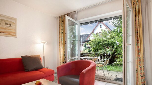 Una habitación con un sofá rojo, un sillón gris, una mesa con fruta y vista a una casa a través de un ventanal.