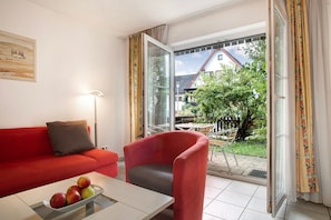 A room with a red sofa, a grey armchair, a table with fruit, and a view of a house through a glass door.