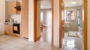 Cocina moderna con armarios de madera, lavavajillas y horno. Hay un comedor con sillas y una mesa. El baño tiene una ducha con cortina.
