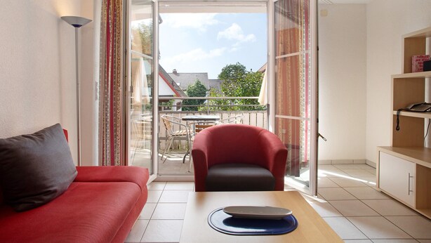 Una habitación con un sofá rojo, una mesa con un ordenador portátil, una silla roja, una estantería y un ventanal corredero que da a una zona exterior con una mesa y sillas.