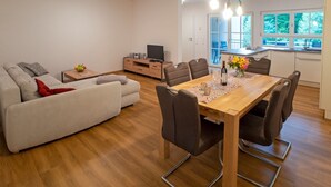 Una sala de estar moderna con sofá, mesa de comedor y sillas. Hay una zona de cocina con ventana, un televisor y una mesita con un jarrón de flores.
