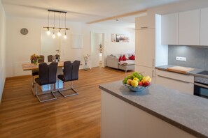 A modern kitchen with a fruit bowl, a dining area with chairs and a table, and a living area with a sofa and a painting.