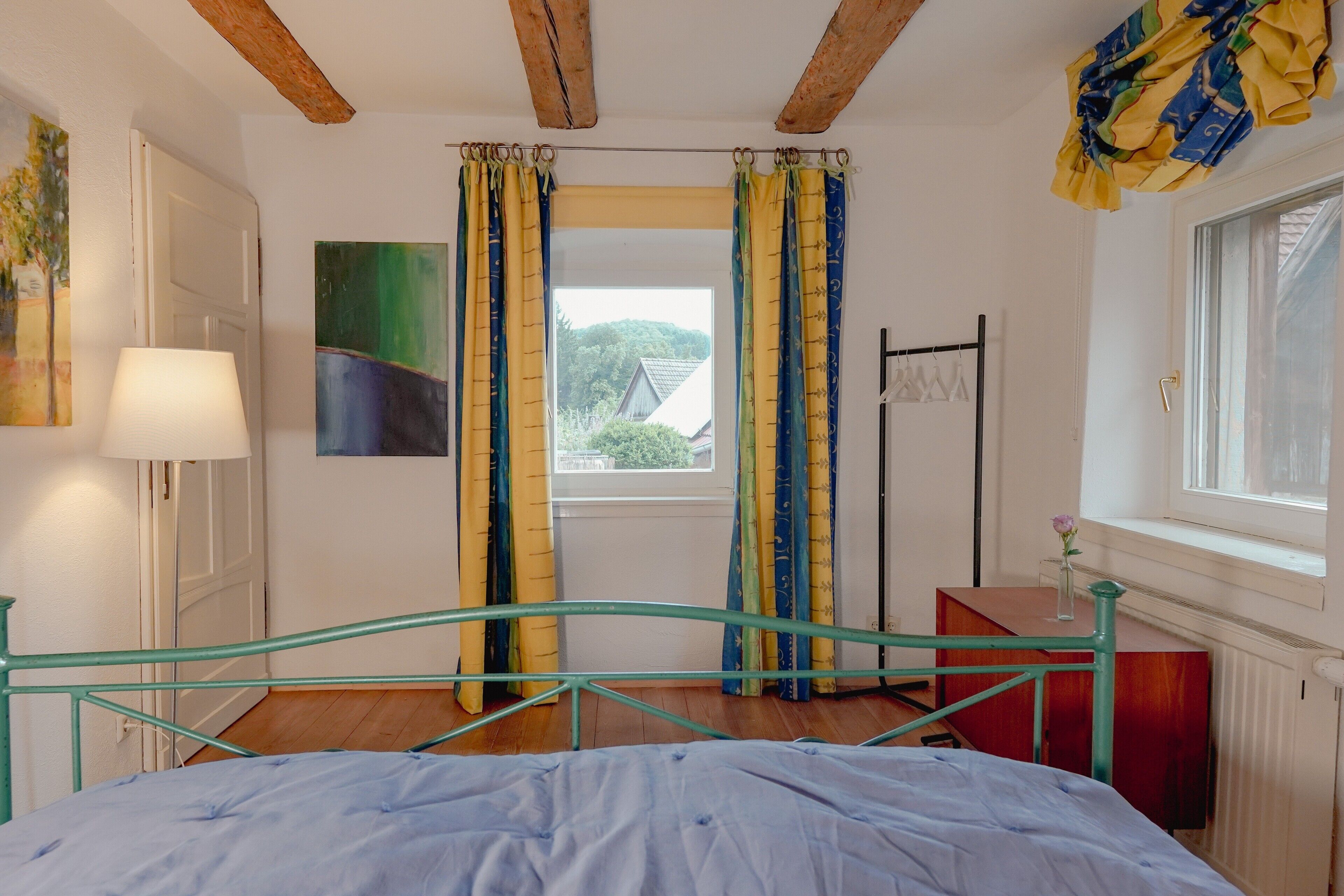 Un dormitorio con cama, ventana con cortinas, lámpara y un cuadro en la pared.