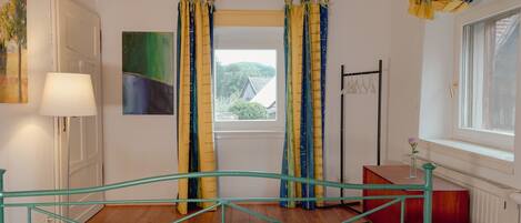 Un dormitorio con cama, ventana con cortinas, lámpara y un cuadro en la pared.