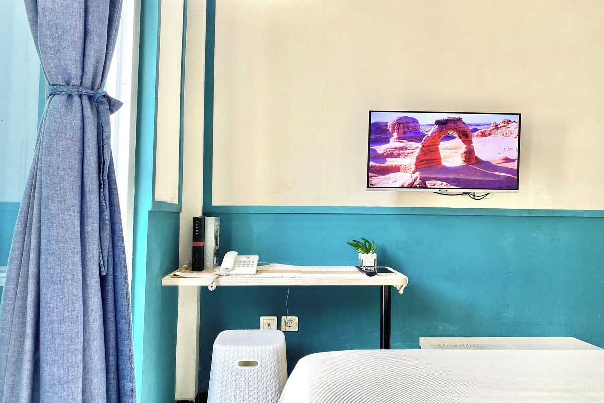 A room with a teal wall, a mounted TV displaying a rock formation, a small desk with a phone and a plant, and a white bed.