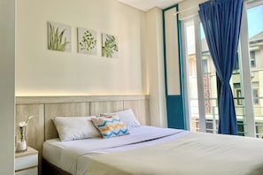 A neatly made bed with white bedding and a blue curtain, a bedside table with a vase, and wall art depicting plants.