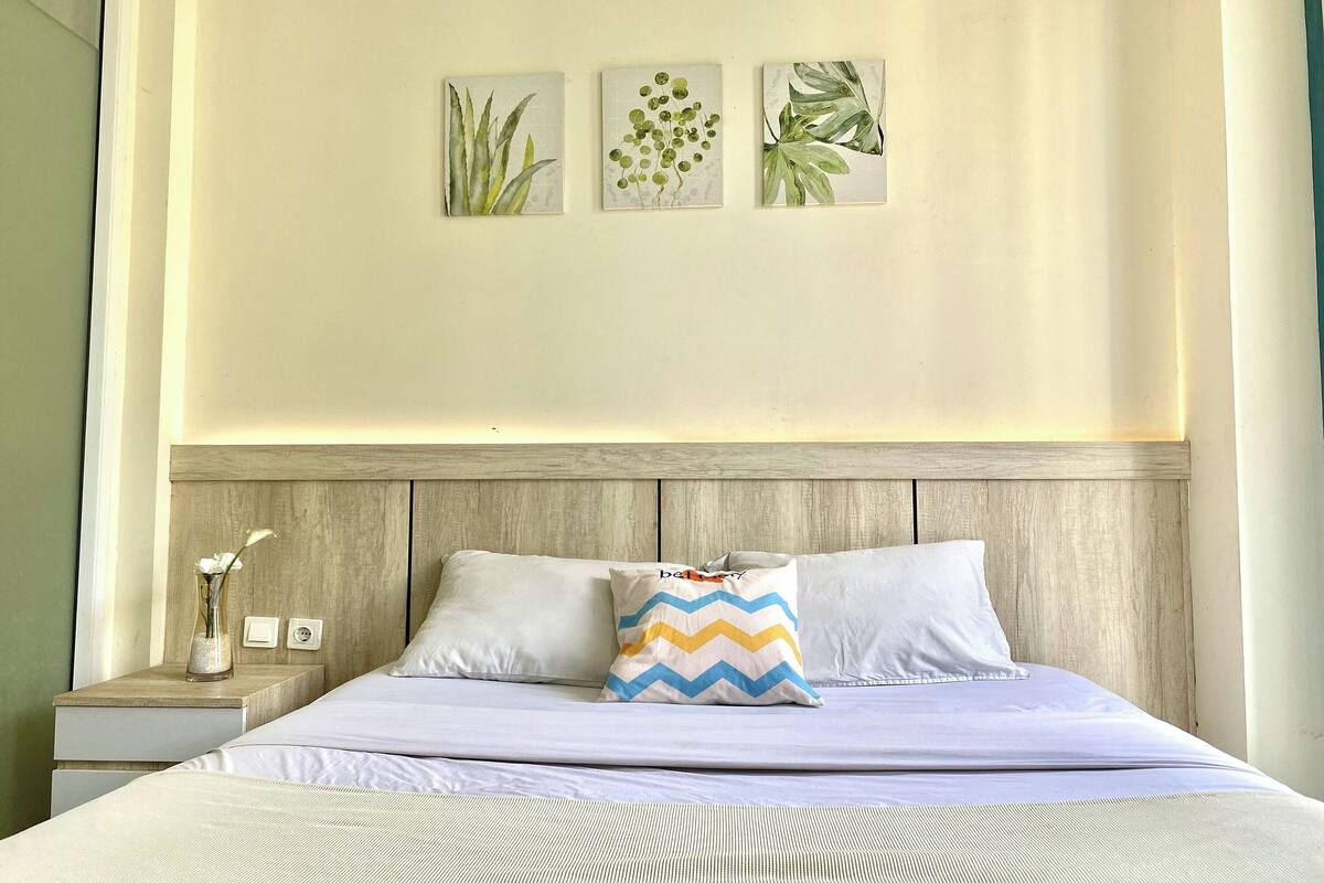 A neatly made bed with a white and blue zigzag pillow, a wooden headboard, and three framed botanical prints on the wall.