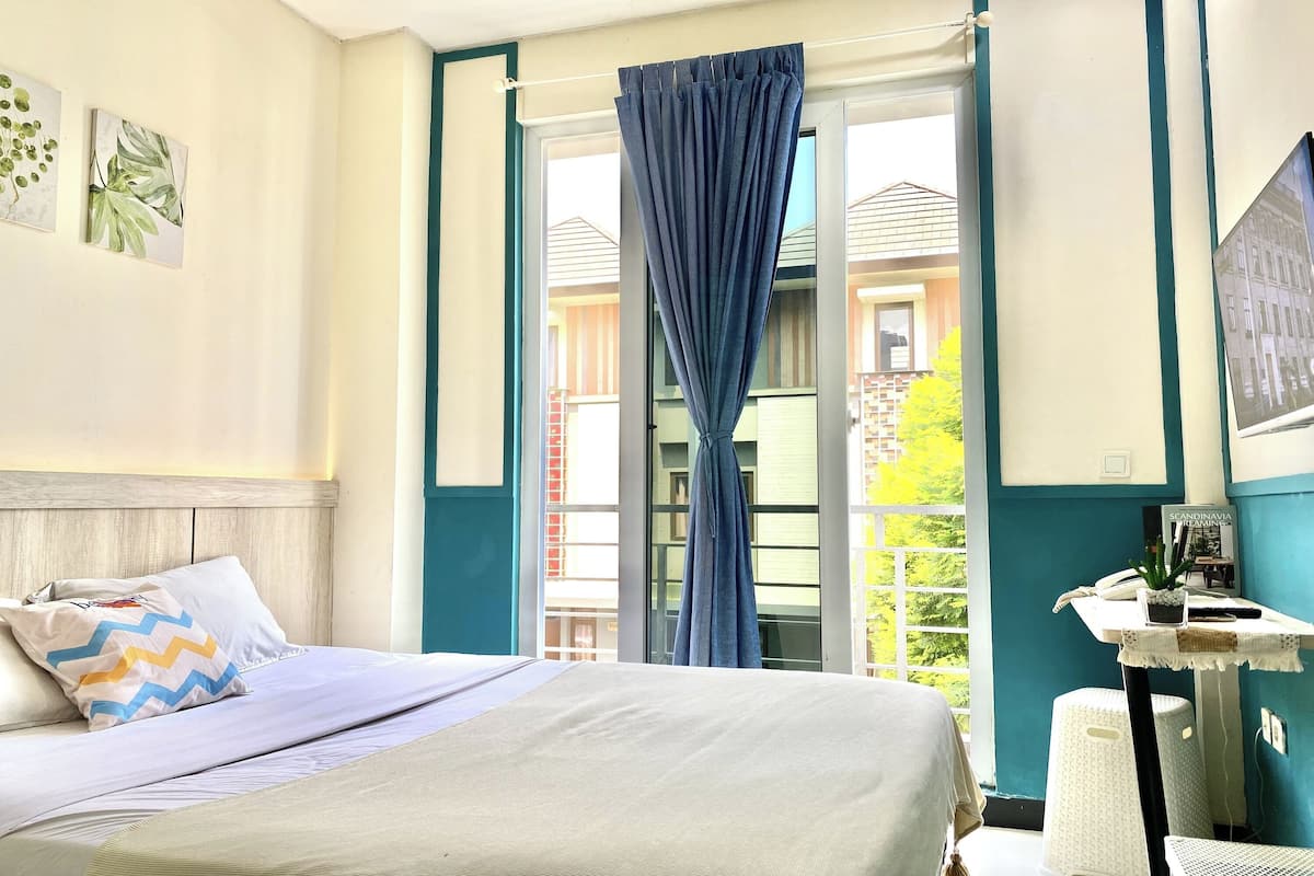 A bedroom with a bed, a window with curtains, a small table with a plant, and a view of buildings outside.